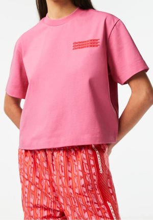 Kobieta ubrana w różowy krótki top z krótkim rękawem z małym czerwonym napisem i czerwone spodnie w wzory, stoi z rękami za plecami.