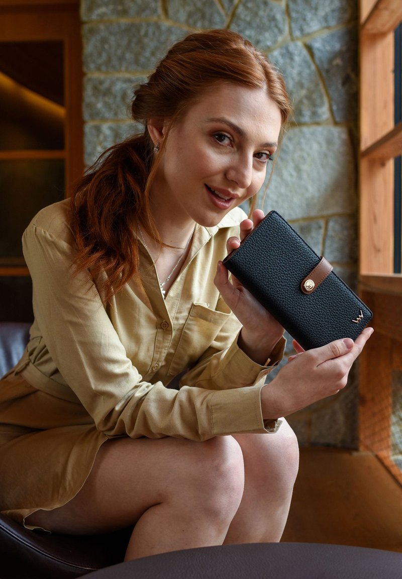 Black textured wallet with a brown leather strap and gold hardware. Woman holds it against a stone wall, wearing a beige collared shirt.