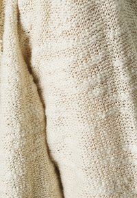 Krämfärgad stickad tyg med ett varierat mönster och subtil lyster, med lösa, öppna stygn och en mjuk, taktil yta.