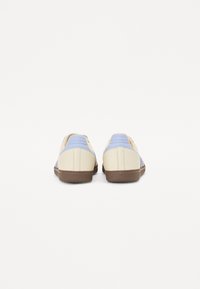 Ett par krämfärgade och ljusblå sneakers med bruna sulor visade från baksidan på en vit bakgrund.