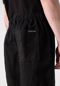 Černé šortky vyrobené z lehkého materiálu, s elastickým pasem, boční kapsou a malou štítkovou značkou Calvin Klein.