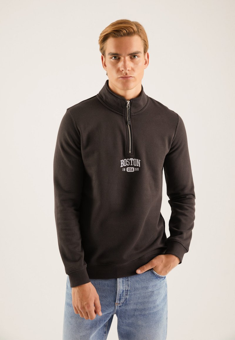 Sudadera negra con media cremallera y cuello alto, con un logo impreso en blanco que dice "BOSTON 1998" centrado en el pecho. Textura de tela suave.