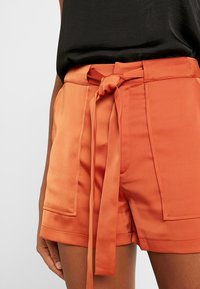 Pomarańczowe, wysokowalne krótkie spodenki z paskiem do wiązania, dwoma przednimi kieszeniami oraz gładkim, teksturowanym wykończeniem.