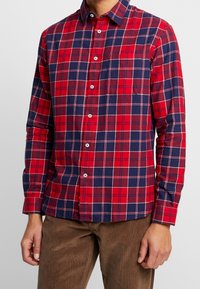 Mężczyzna ubrany w dopasowaną czerwoną i granatową koszulę w kratę na guziki z białymi guzikami, w zestawie z brązowymi sztruksowymi spodniami, stojący z rozluźnionymi rękami.