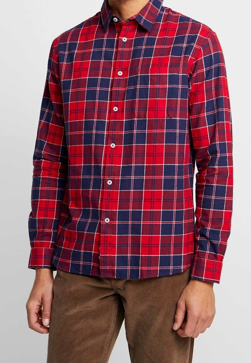 Mężczyzna ubrany w dopasowaną czerwoną i granatową koszulę w kratę na guziki z białymi guzikami, w zestawie z brązowymi sztruksowymi spodniami, stojący z rozluźnionymi rękami.
