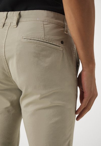 Beige trousers with a smooth texture, featuring back pocket with a flapped design and a decorative button detail. Classic fit.