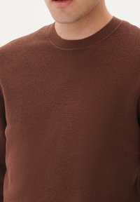 Brown knit sweater with a textured weave, featuring a round neckline and ribbed collar. Focus on the chest area, showing fabric detail.