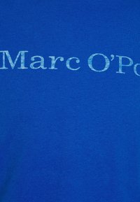 Tissu bleu avec un texte blanc délavé lisant "Marc O'P", partiellement visible au centre.