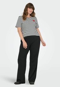 Stripete t-skjorte med kort erme i hvit og svart med rød detalj, parret med løse svarte bukser, iført mørke sko.