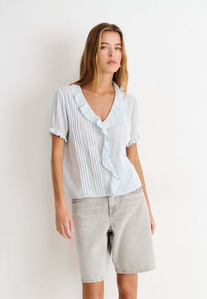 Jeune femme portant un chemisier bleu clair à volants et un short en jean gris clair longueur genou, debout sur fond blanc.