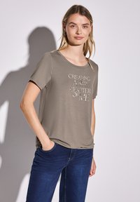 Camiseta de manga corta verde oliva con cuello redondo; presenta texto plateado que dice "CREANDO UN NUEVO CAPÍTULO DE MÍ." Combinada con pantalones vaqueros azules.