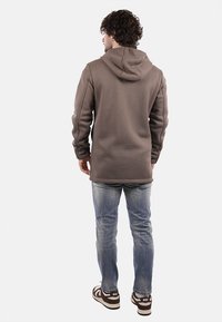 Felpa marrone, con texture, dotata di tasca a marsupio e cappuccio con cordini, abbinata a jeans chiari, strappati e scarpe sportive marroni.