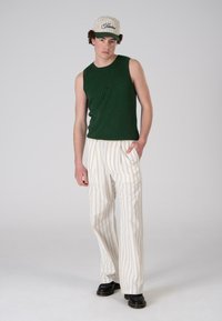 Green ribbed tank top, white striped wide-leg pants, black boots, and a cream cap with green accents and logo on the front.