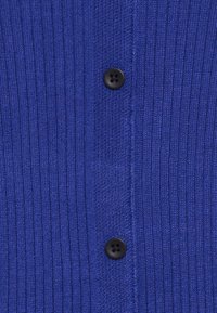 Djupt blå ribbad stickad kofta med en strukturerad yta och svart fyrhåls knappstängning. Tyget verkar mjukt och elastiskt.