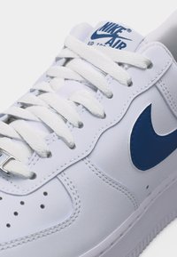 Λευκό αθλητικό παπούτσι Nike Air Force 1 με λευκά κορδόνια και μπλε λογότυπο Nike στη πλευρά, με λεπτομερείς ραφές και οπές.