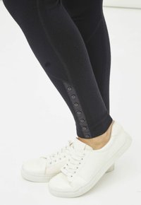 Leggings negros con un acabado texturizado, con un detalle de botón lateral, combinados con zapatillas blancas y suela lisa.