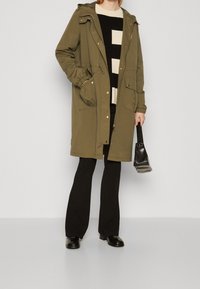 Olivgrüne Parka-Jacke mit Kapuze, Reißverschluss und zwei Fronttaschen, kombiniert mit einem schwarz-weiß gestreiften Pullover und schwarzen Hosen.