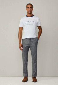 Muž v bielom tričku "Hackett London", šedých prúžkovaných nohaviciach a hnedých formálnych topánkach, stojí s jednou rukou v vrecku.