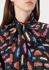 Svart bluse med et mønster av fargerike biler i rødt, blått, rosa og oransje. Stoffet er glatt, og den har et bånd ved halsen.