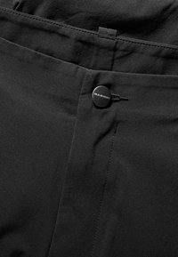 Close-up van zwarte stof met een knoop waarop "Mammut" staat, op een tailleband met zichtbare stiksels en een kleine lus voor een riem.