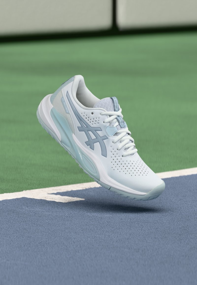 Scarpa da ginnastica azzurro chiaro con tomaia traforata, lacci e tallone di supporto. Presenta un logo decorativo e una suola testurizzata su una superficie da campo.