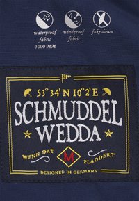 Marinfärgad tygpatch med vit och gul broderi. Texten inkluderar "SCHMUDDEL WEDDA" samt ikoner för vattentåliga, vindtåliga och syntetiskt dun egenskaper.