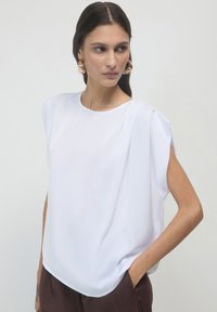Blusa bianca a maniche corte con una vestibilità morbida, caratterizzata da morbide pieghe e una texture liscia, abbinata a pantaloni marroni, mani nelle tasche.