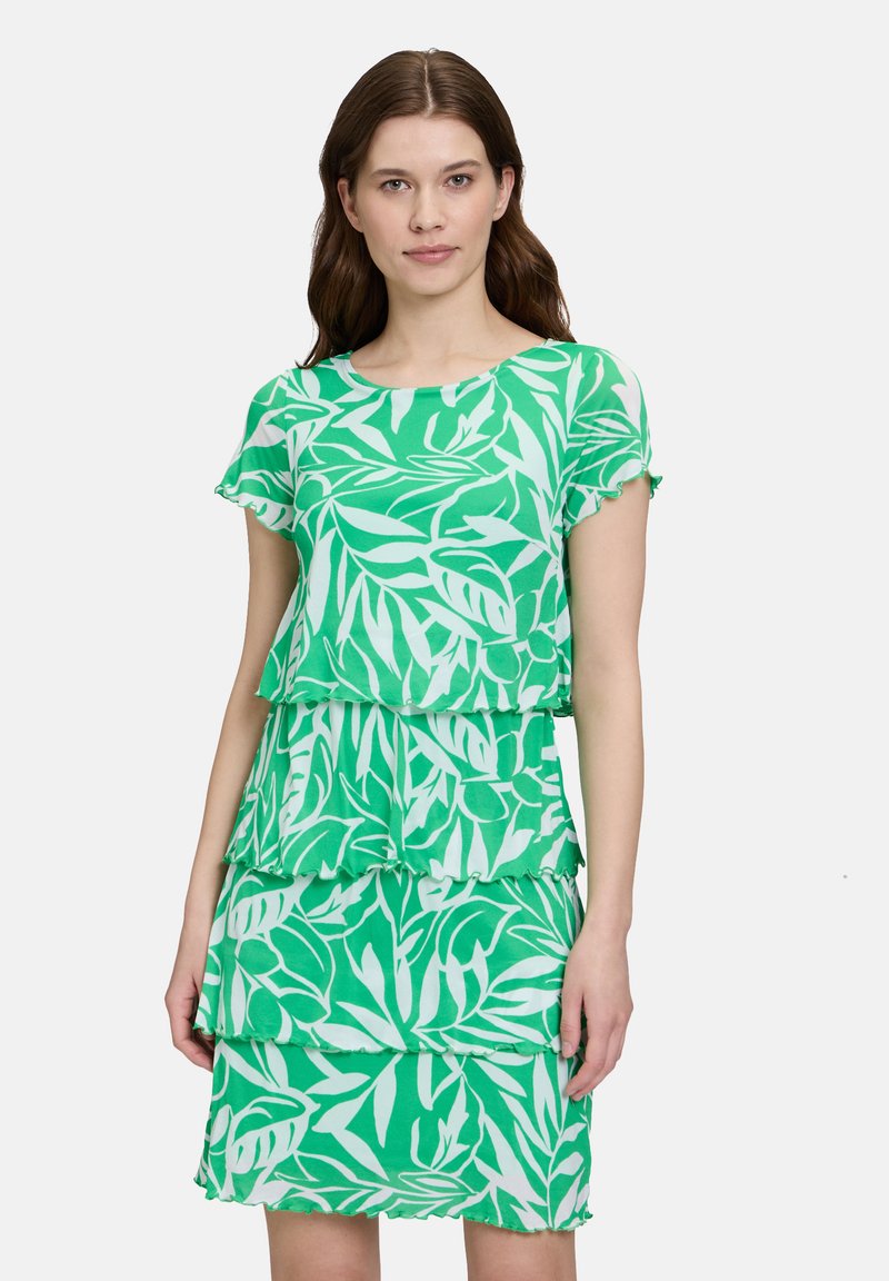 Das grüne, geschichtete Kleid weist ein Blattmuster, kurze Ärmel und einen runden Ausschnitt auf. Der Stoff hat eine glatte Textur mit gerüschten Kanten.