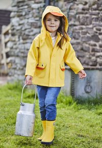 Impermeable amarillo con temática de patos en los bolsillos, ojos azules, combinado con jeans azules y botas de goma amarillas, sosteniendo una regadera de metal.