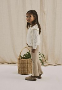 Maglione bianco lavorato a maglia con maniche arricciate, pantaloni beige a coste e scarpe marroni senza lacci; tiene in mano un cesto intrecciato con del verde.