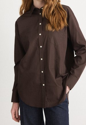 Personne portant une chemise marron foncé boutonnée avec une poche et un jean bleu, main dans la poche, sur un fond clair uni.