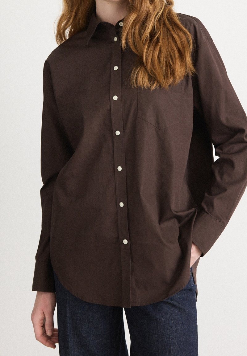 Personne portant une chemise marron foncé boutonnée avec une poche et un jean bleu, main dans la poche, sur un fond clair uni.