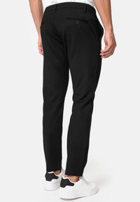 Zwarte, op maat gemaakte broek met een slanke pasvorm, voorzien van een achterzak met knoopdetail, gecombineerd met witte sneakers. Gladde stoftextuur.