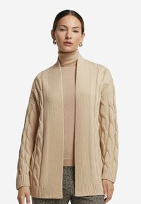 Beige kofta med flätat mönster och ribbad kant, öppen framsida, kombinerad med en ljusbrun polotröja och mönstrade byxor.