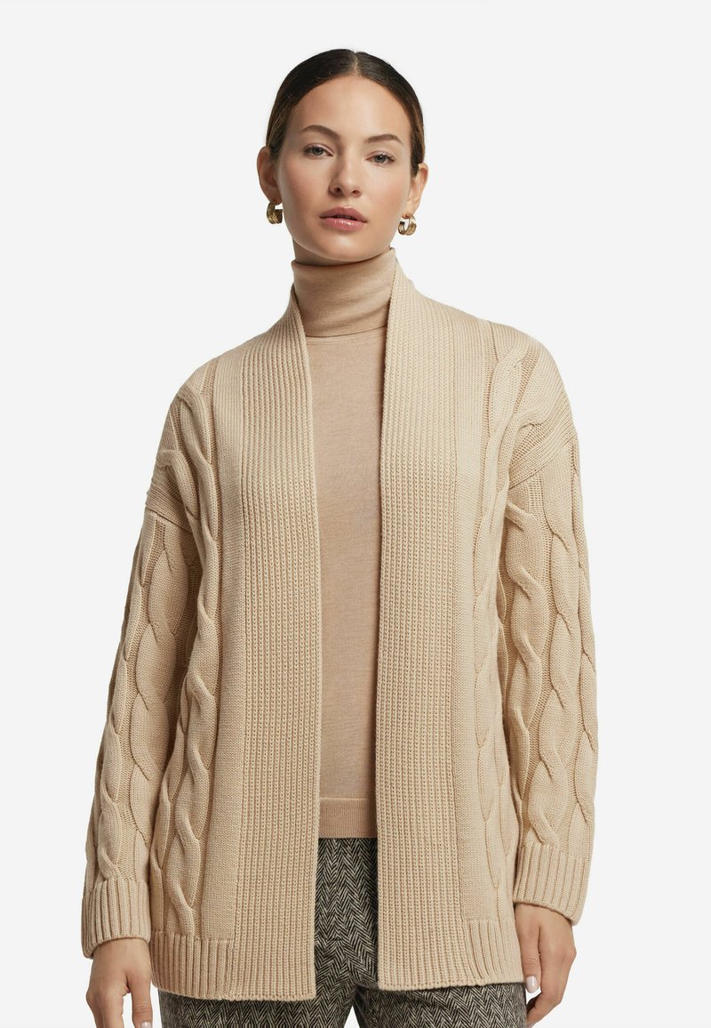 Beige kofta med flätat mönster och ribbad kant, öppen framsida, kombinerad med en ljusbrun polotröja och mönstrade byxor.