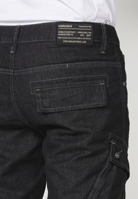 Schwarze Jeans-Cargohosen mit mehreren Taschen, einem silbernen Knopf und Kontrastnähten. Auf der Rückseite befindet sich ein Markenetikett mit Textdetails.