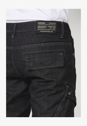 Schwarze Jeans-Cargohosen mit mehreren Taschen, einem silbernen Knopf und Kontrastnähten. Auf der Rückseite befindet sich ein Markenetikett mit Textdetails.