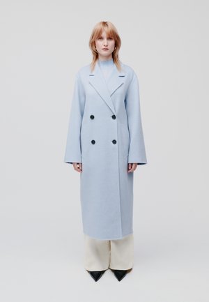 Hellblauer, doppelt gehängter Mantel aus weichem Material, mit großen schwarzen Knöpfen und weiten Ärmeln, kombiniert mit cremefarbenen Hosen und spitzen schwarzen Schuhen.