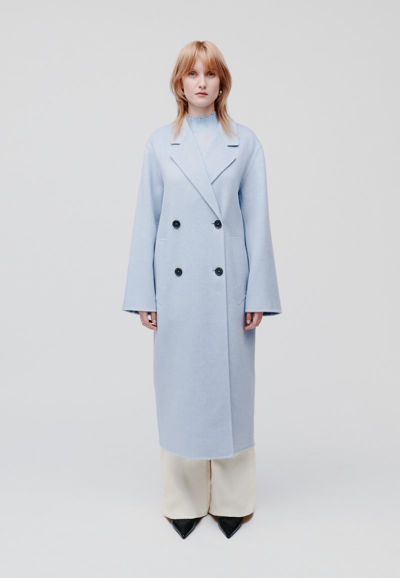 Cappotto doppiopetto azzurro chiaro in morbido tessuto, con grandi bottoni neri e maniche ampie, indossato sopra pantaloni crema e scarpe nere a punta.