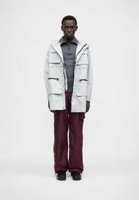 Helehall veekindel jope, millel on mitmed esikotid, koos burgundi värvi cargo pükste ja külgsiribadega ning mustade jalatsitega.