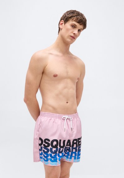 Άνδρας χωρίς πουκάμισο που φοράει ροζ μαγιό με μαύρο και μπλε λογότυπο "DSQUARED", στέκεται μπροστά σε απλό φωτεινό φόντο.