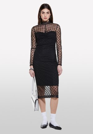 Robe noire avec des manches transparentes à pois et un corps texturé, décolleté en forme de scoop et ourlet au genou. Associée à des chaussettes blanches et des ballerines noires.