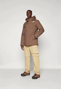 Vyras vilki rudą izoliacinę parka su pūkų apdaila ant kapučino, kreminės spalvos krovinių kelnes ir rudus žygių batus, stovintis prieš paprastą foną.