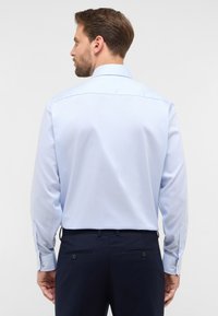 Camicia a maniche lunghe azzurro chiaro con sottili righe bianche, dotata di colletto classico e polsini con button, indossata con pantaloni scuri su misura.