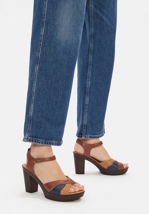 Sandały na obcasie w kolorach brązowym i niebieskim z paskiem wokół kostki, z kwadratowym noskiem i masywnym obcasem. Noszone z dżinsowymi spodniami.