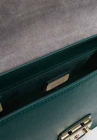 Interiér smaragdově zelené kožené kabelky s kapsou na zip, zlatými akcenty a označením značky. Texturovaný povrch a strukturovaný tvar.