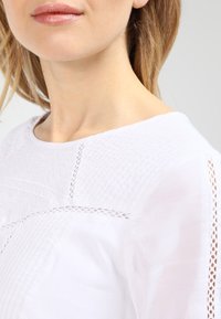 Blouse blanche avec des détails en dentelle complexe et un tissu texturé, présentant un col rond et des accents ajourés sur les manches.