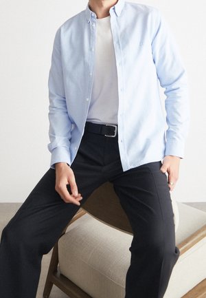 Mężczyzna siedzący na beżowym, drewnianym krześle z poduszką, ubrany w jasnoniebieską koszulę z guzikami na białym tle oraz czarne spodnie z czarnym paskiem.