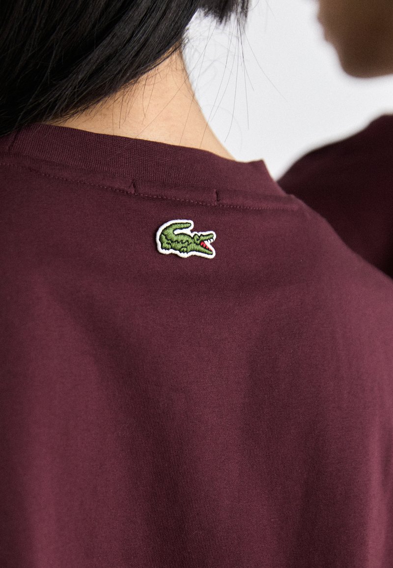 T-shirt en coton bordeaux avec un logo de crocodile brodé vert au dos, près de l'encolure. Texture lisse, coupe décontractée.