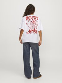 Camiseta blanca de gran tamaño con estampado gráfico rojo que dice "HOWDY" y varias ilustraciones con temática del oeste en la parte trasera. Se lleva con pantalones oscuros de mezclilla.